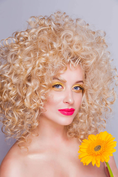 Belle fille blonde à la mode dans un style rétro avec une coiffure bouclée volumineuse, les épaules ouvertes et un tournesol dans ses mains sur un fond gris
.  - Photo, image