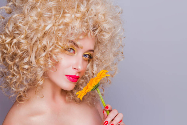 Beautiful fashionable blonde girl in retro style with voluminous curly hairstyle, open shoulders and a sunflower in her hands on a gray background.  - Zdjęcie, obraz