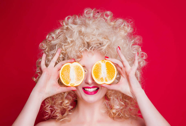 Studio portrait of Beautiful fashionable blonde girl in retro style with voluminous curly hairstyle, open shoulders,  with oranges covering eyes on red background - Fotografie, Obrázek