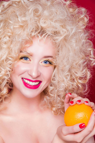 schöne modische blonde Mädchen im Retro-Stil mit voluminöser Lockenfrisur, nackten Schultern und einer Orange in ihren Händen blickt in die Kamera und lächelt - Foto, Bild
