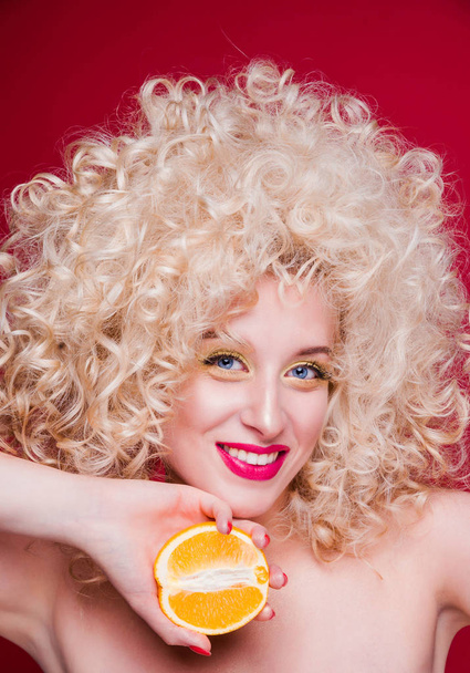 Beautiful fashionable blonde girl in retro style with voluminous curly hairstyle, bare shoulders and an orange in her hands looks at the camera and smiles - Zdjęcie, obraz