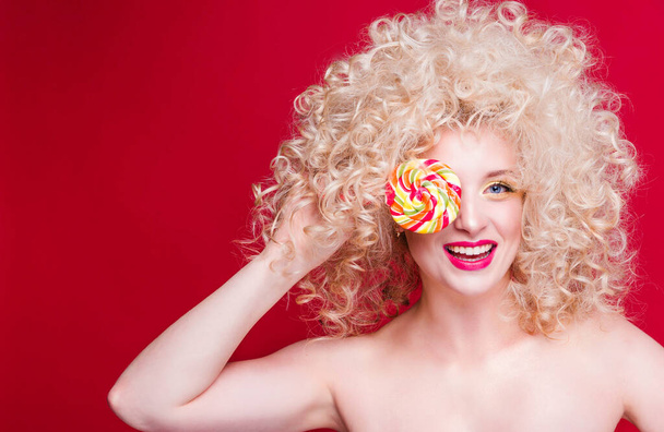 Beautiful fashionable blonde girl in retro style with voluminous curly hairstyle, open shoulders on a red background smiles, laughs and closes one eye with colored candy - Valokuva, kuva