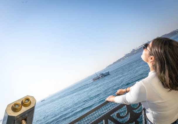 Mladá brunetka modelka pózuje s hlavou opřenou dozadu a rukama na kovovém plotu s mořem, lodí a přístavem v pozadí. Istanbul. Turistická destinace. Prázdné místo. - Fotografie, Obrázek