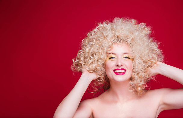 Schöne modische Blondine im Retro-Stil mit voluminöser Lockenfrisur, offenen Schultern auf rotem Hintergrund blickt freudig in die Kamera und berührt die Frisur mit den Händen - Foto, Bild