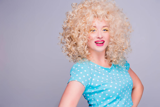 Schöne Blondine im Retro-Stil mit voluminöser Lockenfrisur, in einer blauen Tupfen-Bluse auf grauem Hintergrund, lächelt und blickt in die Kamera - Foto, Bild