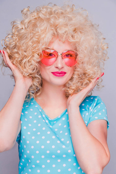 Belle fille blonde de style rétro avec une coiffure bouclée volumineuse, dans un chemisier à pois bleus et des lunettes roses sur un fond gris
 - Photo, image