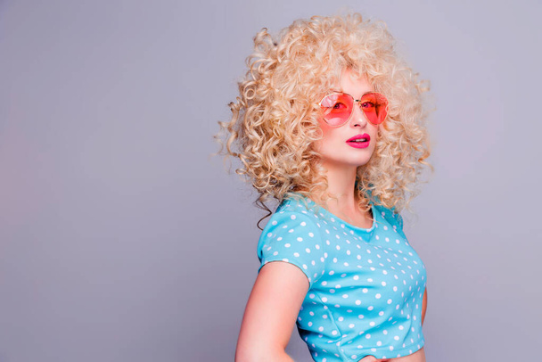 Piękna blondynka w stylu retro z obszerną kręconą fryzurą, w niebieskiej bluzce w kropki i różowych okularach na szarym tle - Zdjęcie, obraz