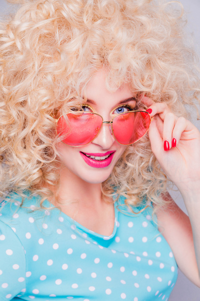 Kaunis retro-tyylinen blondi tyttö, jolla on laaja kihara kampaus, sinisessä polka-dot-puserossa ja vaaleanpunaiset lasit harmaalla taustalla
 - Valokuva, kuva