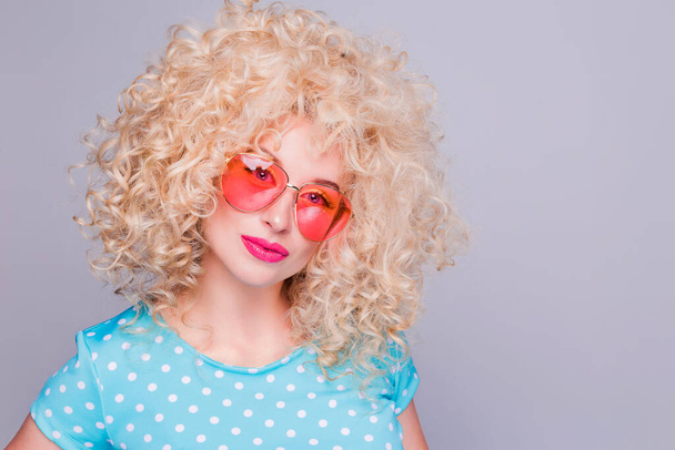 Beautiful retro-style blonde girl with voluminous curly hairstyle, in a blue polka-dot blouse and pink glasses on a gray background - Photo, Image