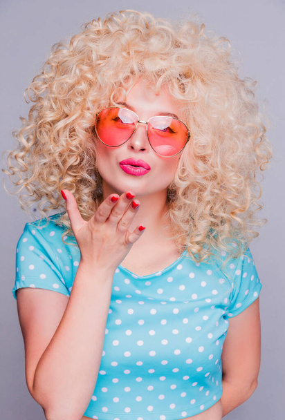 Mooi blond meisje in retro stijl met een volumineus krullend kapsel, in een blauwe polka-dot blouse en roze bril op een grijze achtergrond, kijkt naar de camera en stuurt een luchtkus - Foto, afbeelding