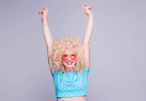 Красивая блондинка в стиле ретро с объемной кудрявой прической в голубой блузке в горошек и розовых очках на сером фоне испытывает радость и восторг
 - Фото, изображение