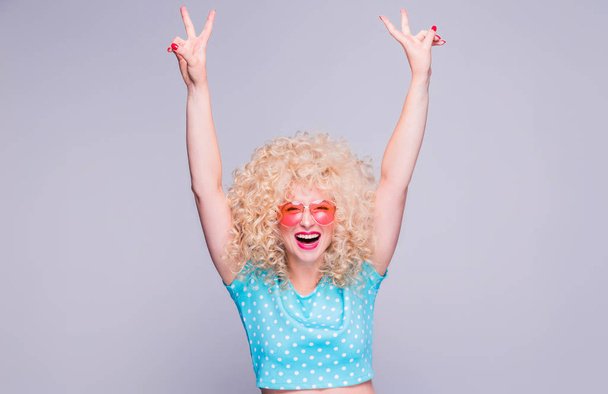Beautiful blonde girl in retro style with voluminous curly hairstyle in a blue polka-dot blouse and pink glasses on a gray background experiences joy and delight - Foto, Bild