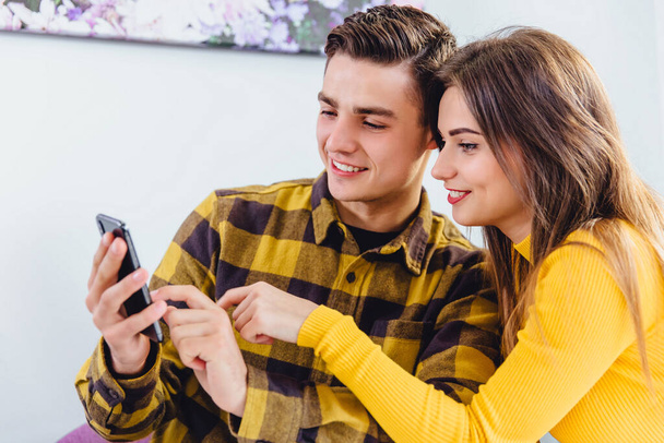 Lively young couple looking through photos on smartphone, smiling with pleasure as they relax at home. - Fotografie, Obrázek