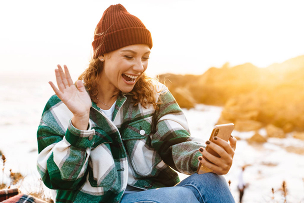 Image of excited woman using smartphone and waving hand while sitting - Фото, зображення