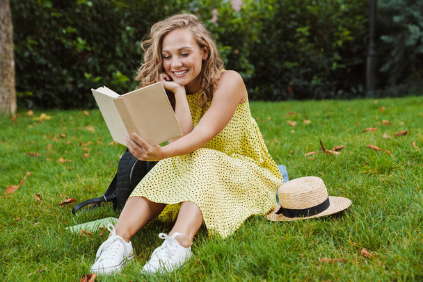 Αισιόδοξη γυναίκα κάθεται στο γρασίδι διαβάζοντας το βιβλίο. - Φωτογραφία, εικόνα
