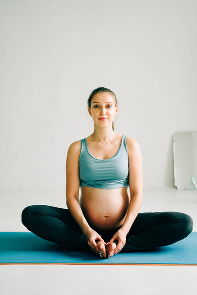 una joven bastante embarazada en ropa deportiva está haciendo yoga, haciendo asana Baddha Konasana en la alfombra en el estudio
 - Foto, Imagen