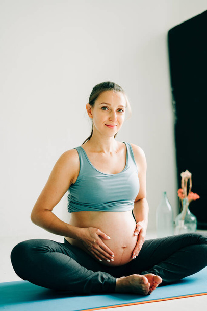 a young pretty pregnant girl in sports clothes is doing yoga, doing asana Baddha Konasana on the Mat in the Studio - 写真・画像