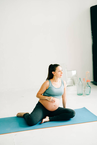 beautiful pregnant girl in sportswear sitting on a yoga Mat and stroking her belly on a studio - 写真・画像
