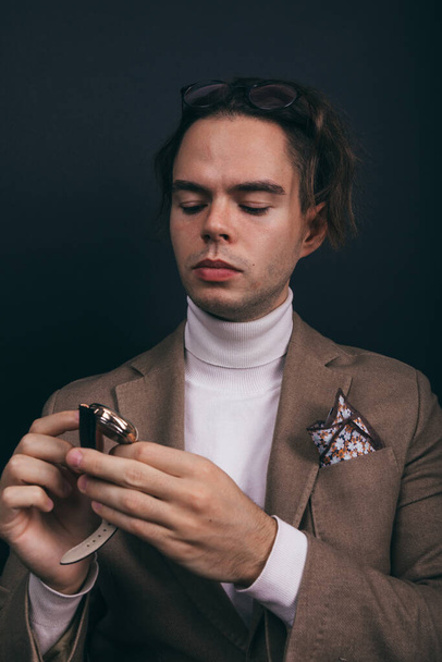 A dapper young man obviously you wearing a white turtleneck sweater and a beige jacket with handkerchief in his top breast pocket, looking pensively at his wrist watch, contemplating time - 写真・画像
