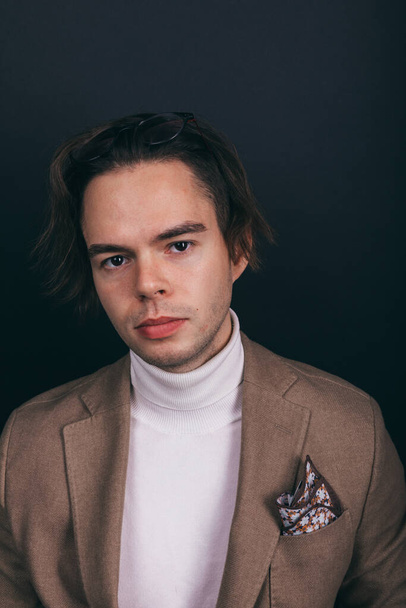A dapper young man obviously you wearing a white turtleneck sweater and a beige jacket with handkerchief in his top breast pocket, looking pensively at his wrist watch, contemplating time - Foto, afbeelding