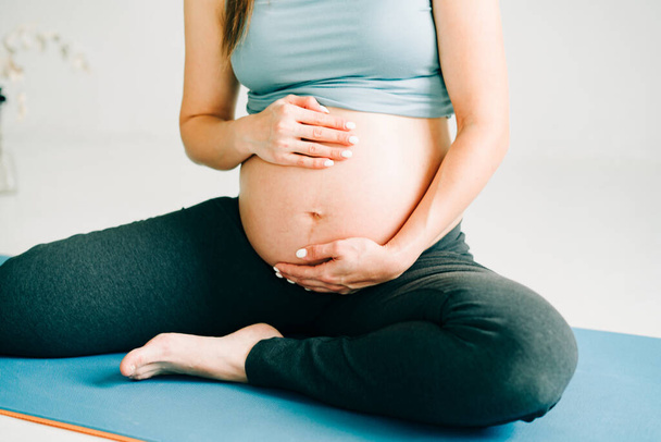 mooi zwanger meisje in sportkleding zitten op een yoga mat en strelen haar buik op een studio - Foto, afbeelding