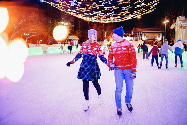 Buz pateni pistinde sıcak kırmızı süveter giyen genç ve güzel aşıklar arka plan aydınlatmalarında beyaz patenler giyiyorlardı. - Fotoğraf, Görsel
