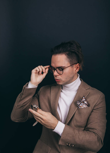 Un jeune homme habillé d'un pull à col roulé blanc et d'une veste beige avec mouchoir dans sa poche supérieure, regardant pensivement sa montre-bracelet, contemplant le temps
 - Photo, image
