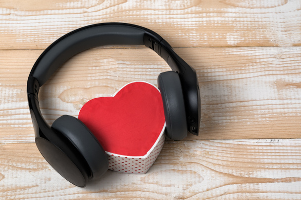 Full-size wireless headphones pulled over small red heart-shaped box on a light brown wooden table. Love music concept - Photo, Image