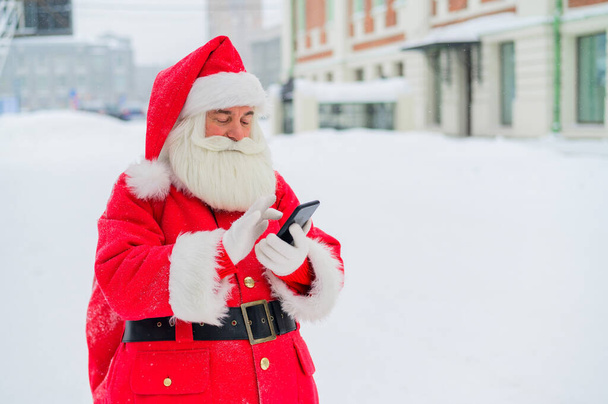 Surprised santa claus is using mobile outside. Santa Claus stands in the background of buildings in winter and calls on smartphone. Snow in December. - Фото, зображення