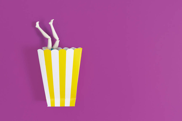 Time for party. Female legs peek out from paper cup for popcorn on a purple background. Copy space for text - Foto, afbeelding