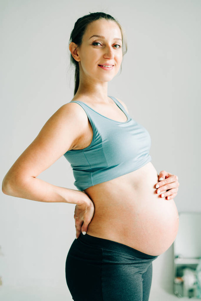uma jovem grávida em sportswear está de pé em um tapete de ioga, sorrindo e segurando sua barriga no estúdio
 - Foto, Imagem