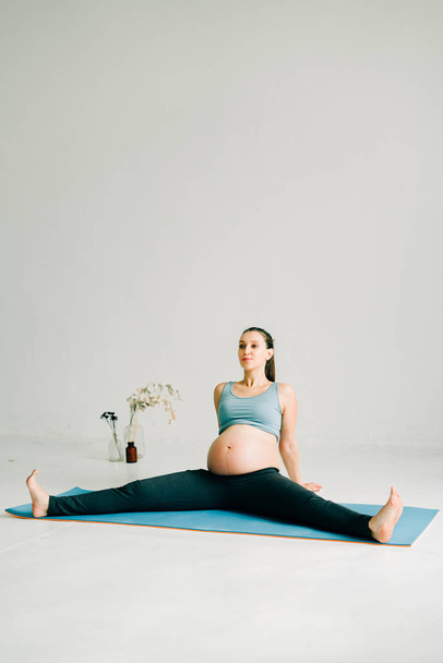 a young beautiful pregnant girl in sports clothes is doing yoga, performing exercises sitting on a rug in the Studio - Foto, Imagem