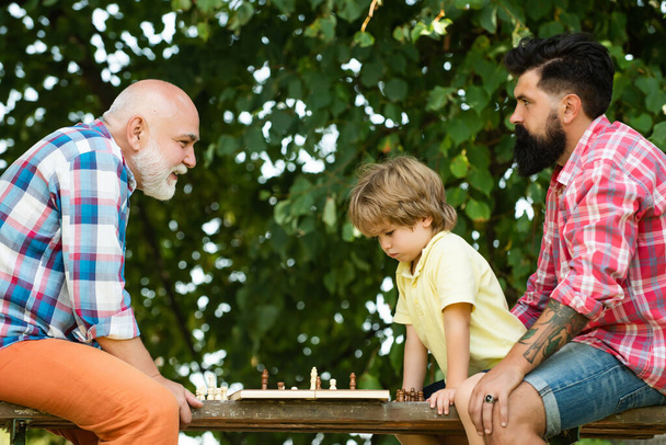 Fathers day. Old man with little boy playing chess. Father and Grandfather with son playing Chess in the park at the day time. - Zdjęcie, obraz