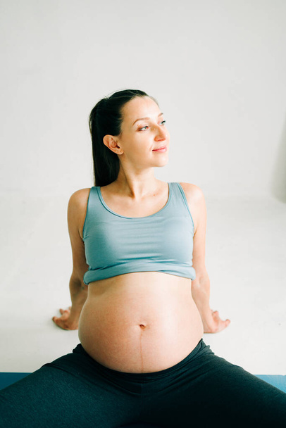 een jong mooi zwanger meisje in sportkleding doet yoga, het uitvoeren van oefeningen zittend op een tapijt in de Studio - Foto, afbeelding