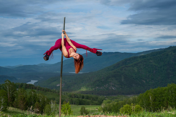 Beautiful red-haired woman dancing on a portable platform on a background of mountains. The girl moves sexually next to the pole. Jumpsuit for classes. high heels. Altai. Awesome flexibility. - Φωτογραφία, εικόνα