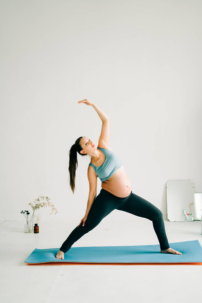 a young pretty pregnant girl in sports clothes is doing yoga, doing asana on the Mat in the Studio - Фото, зображення