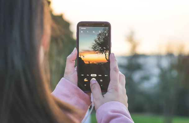 Dłonie kobiet trzymają smartfona i robią zdjęcie zachodu słońca. Fotografia smartfona. - Zdjęcie, obraz