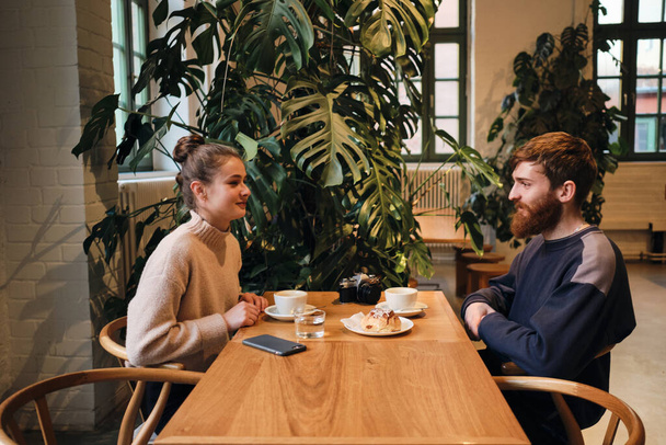Seitenansicht eines jungen schönen Paares, das sich verträumt beim Kaffeetrinken in einem gemütlichen Café ansieht - Foto, Bild