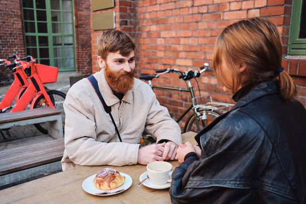 Joven hombre barbudo casual alegremente hablando con su novia descansando juntos en la cafetería de la calle
  - Foto, imagen