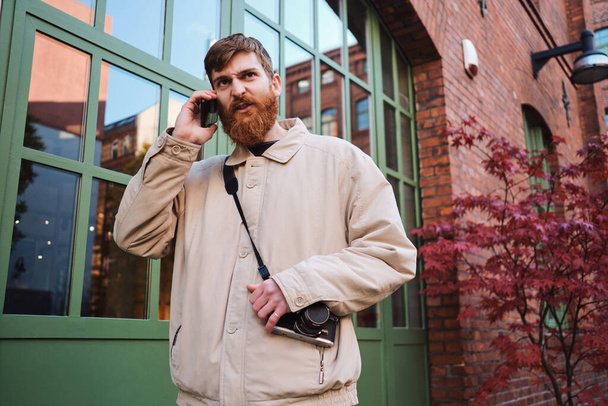 Молодой бородатый мужчина сердито разговаривает по мобильному телефону на открытом воздухе
 - Фото, изображение
