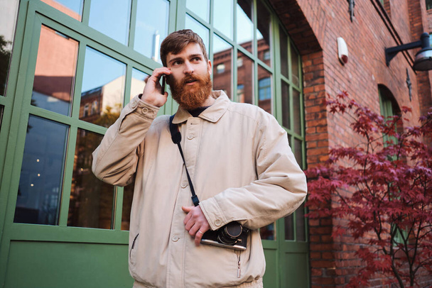 Young casual bearded man intently looking away talking on cellphone outdoor - 写真・画像