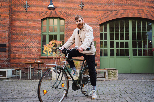 Young attractive bearded man thoughtfully looking in camera with old classic bicycle on street - Photo, Image