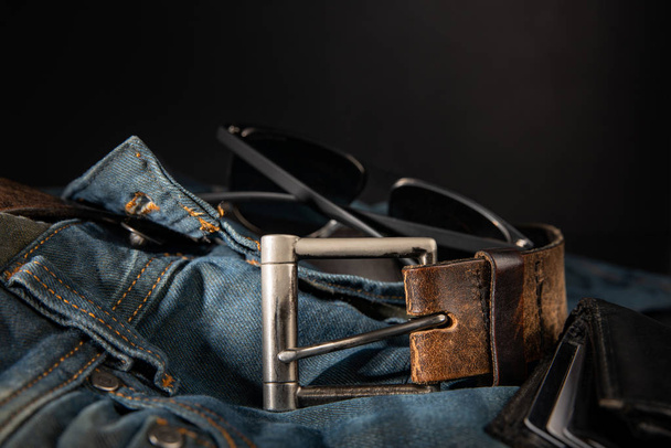 Mens accessories, fashion trends, jeans, a leather belt, an ajar wallet, accidentally lie on a black background with blurred depth of field, close-up. Emphasizes the masculinity of a real man. - Photo, Image