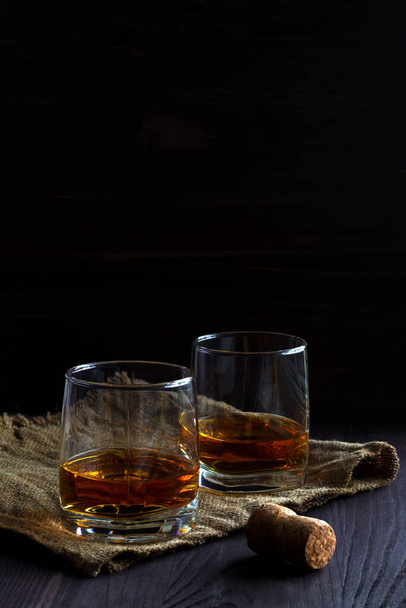 alkoholické nápoje. sklenice whisky s ledem na černém pozadí. dvě sklenice whisky na dřevěném stole s lopatkou a korkem. - Fotografie, Obrázek