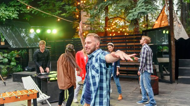 Handsome 30s caucasian young man dancing on near the camera on the background of his friends at the party in cozy evening garden. - Photo, image