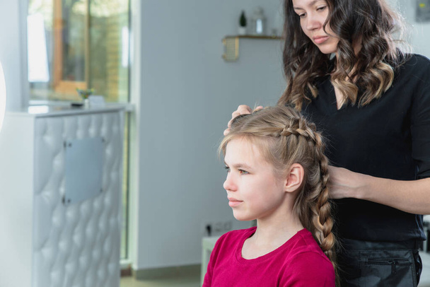 Hairdresser weaves a braid to a preteen blond girl in a beauty and hair salon - Fotoğraf, Görsel