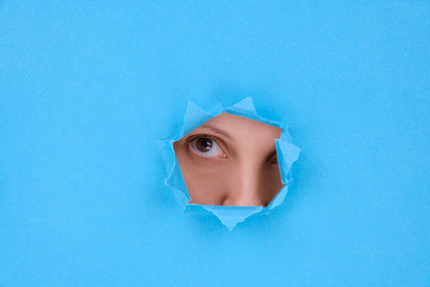 Part of a womans face the eyes and nose of an attractive woman looking into camera, behind a broken wall or torn paper, on blue background. Space for text, banner letterhead, or feminist design - Photo, image