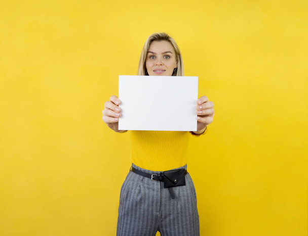 Prachtige jonge vrouw met een model verticaal wit vel papier met kopieerruimte voor tekst op gele achtergrond - Foto, afbeelding