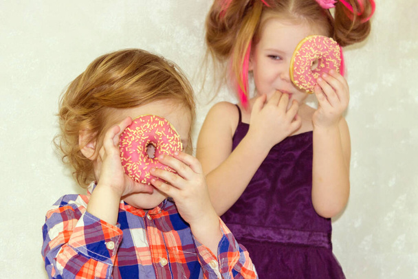 Little happy cute girl is eating donut on white background wall. Child is having fun with donut. Tasty food for playing kids. Funny time  with sweet food. Bright baby girl with pink hair. - Zdjęcie, obraz