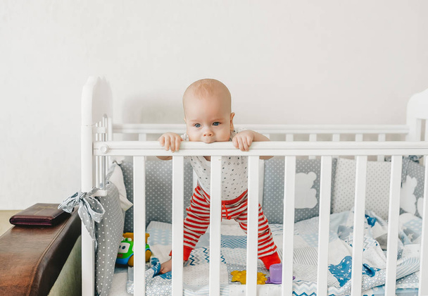 Funny little baby standing in crib at home and tries to taste wooden bed. - Foto, Bild